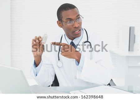Serious male doctor pointing at prescription bottle with laptop in medical office