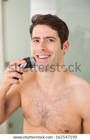 Portrait of a smiling handsome young shirtless man shaving with electric razor