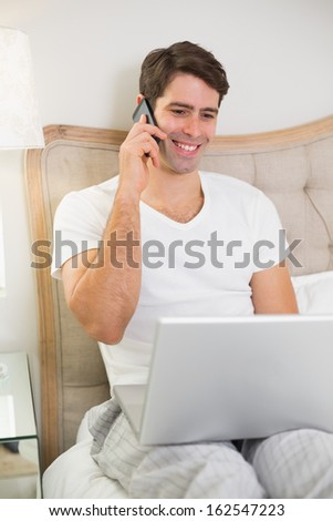 Casual smiling young man using cellphone and laptop in bed at home