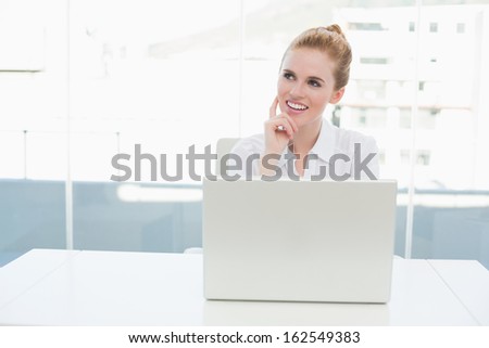 Thoughtful businesswoman looking up while using laptop on desk in a bright office