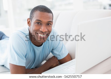 Portrait of a smiling young Afro man with laptop lying on sofa in a bright house