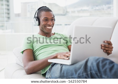 Relaxed smiling young Afro man with headphones using laptop on sofa in a bright house