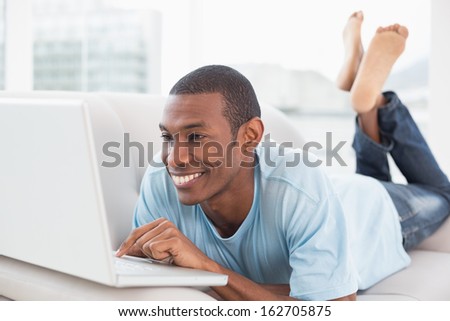 Relaxed casual young Afro man with laptop lying on sofa in a bright house