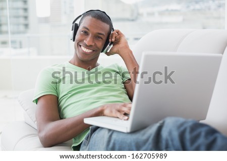 Portrait of a relaxed smiling young Afro man with headphones using laptop on sofa in a bright house