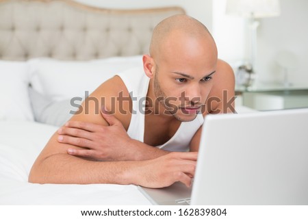Close up of a serious casual bald young man using laptop in bed at home