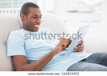 Side view of a casual smiling young Afro man using digital tablet on sofa in a bright house