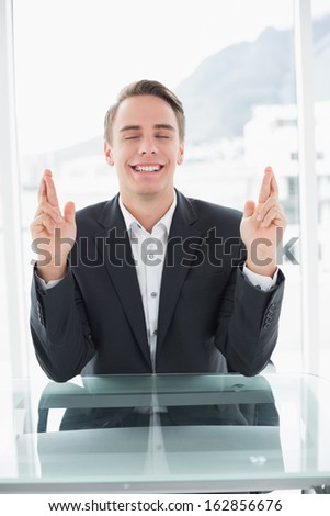 Smiling young businessman with fingers crossed sitting at office desk
