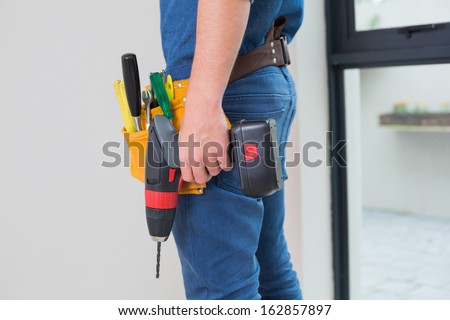 Side view mid section of a handyman with drill and tool belt by the wall