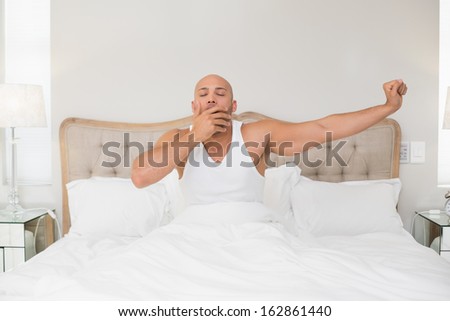 Young bald man waking up in bed and stretching his arms at home