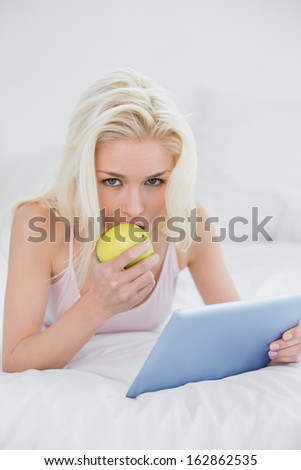 Portrait of a casual young blond with tablet PC eating an apple in bed at home
