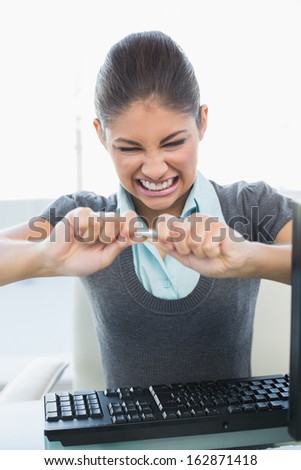Young angry businesswoman breaking pen over computer keyboard in a bright office