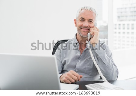 Portrait of a smiling mature businessman on call in front of laptop at desk in a bright office