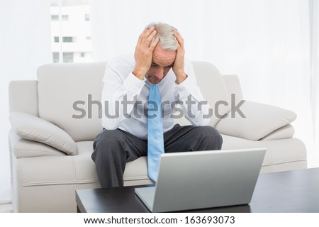 Worried mature businessman with head in hands in front of laptop in the living room at home