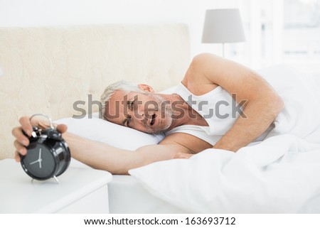 Sleepy mature man extending hand to alarm clock in bed at home