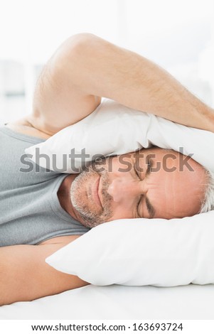 Close-up of a mature sleepy man covering ears with pillow in bed at home