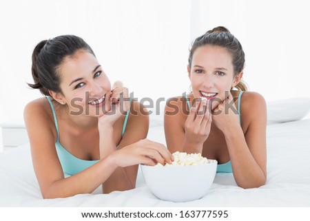 Portrait of two smiling young female friends eating popcorn while lying in bed at home