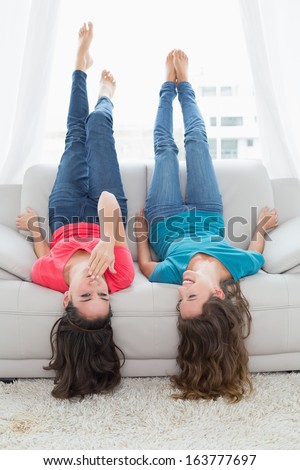 Two smiling young female friends lying on sofa with legs in the air in living room at home