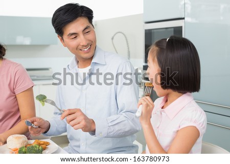 Father watching little girl eating food with a fork in the kitchen at home