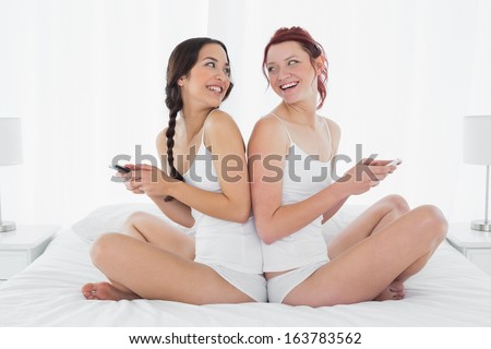 Side view of two happy young female friends sitting back to back with cellphone on bed at home