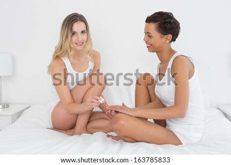 Full length of a pretty young woman painting friend\'s nails on bed at home