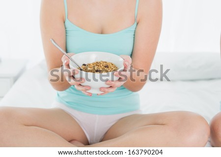 Close-up mid section of a young female with a bowl of cereal sitting on bed at home
