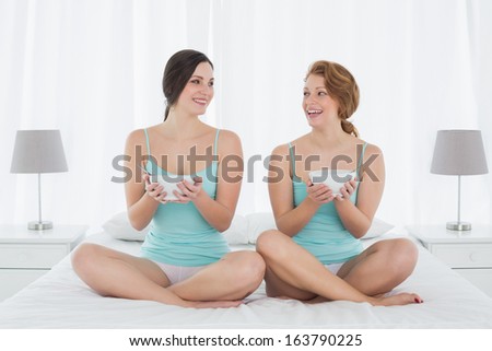 Two smiling young female friends with salad bowls sitting on bed at home