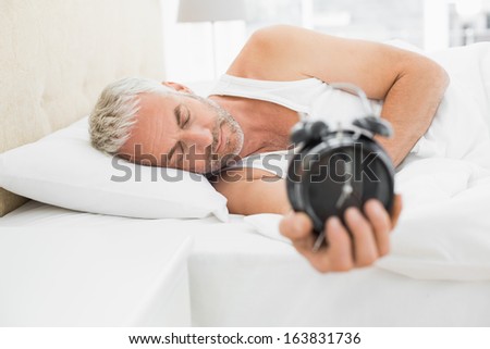 Sleepy mature man holding out alarm clock in bed at home