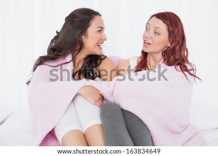 Relaxed young female friends covered in sheet while chatting on bed at home