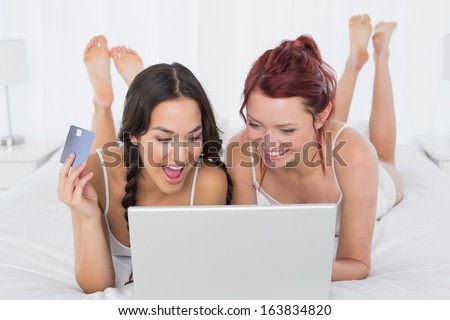 Cheerful relaxed young female friends doing online shopping through laptop and credit card in bed