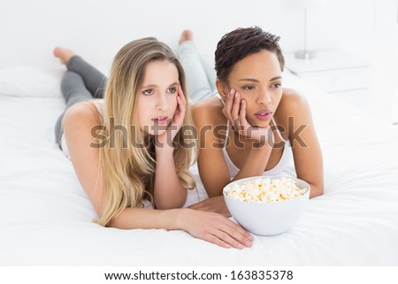 Two serious young female friends with popcorn bowl lying in bed at home