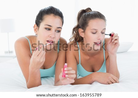 Two young female friends applying make-up while lying in bed at home