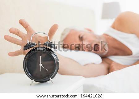 Blurred mature man extending hand to alarm clock in bed at home