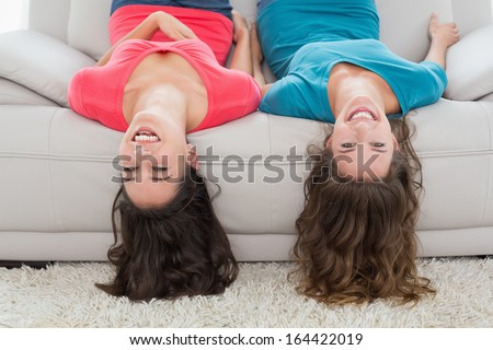 Portrait of two smiling young female friends lying upside down on sofa in living room at home