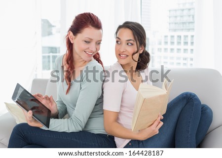 Side view of two relaxed young female friends with digital tablet and book on sofa at home