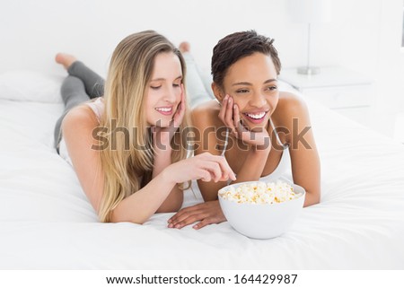 Two cheerful young female friends with popcorn bowl lying in bed at home