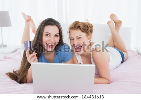 Portrait of cheerful relaxed young female friends  doing online shopping through laptop and credit card in bed