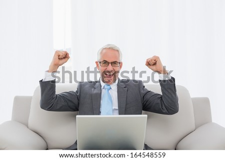 Elegant businessman cheering with clenched fists in front of laptop on sofa in living room at home