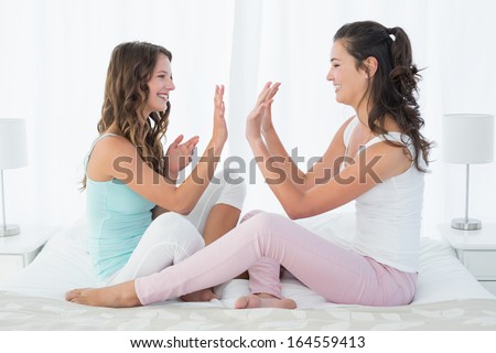 Side view of happy young female friends playing clapping game on bed at home