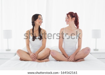 Full length of two happy young female friends in white tank tops sitting on bed at home