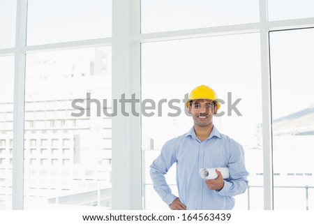 Portrait of a smiling young architect in yellow hard hat with blueprint in a bright office