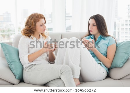 Two relaxed young female friends with coffee cups conversing in the living room at home