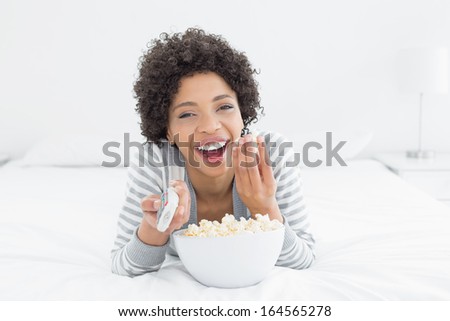 Portrait of a cheerful young woman with remote control and popcorn bowl lying in bed at home