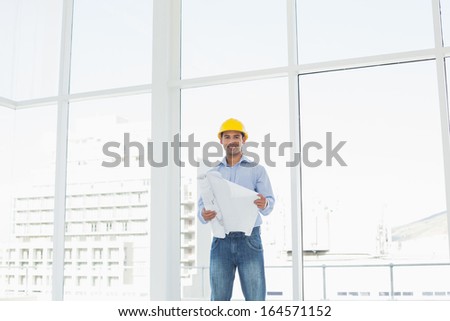 Portrait of a smiling young architect in yellow hard hat with blueprint in a bright office
