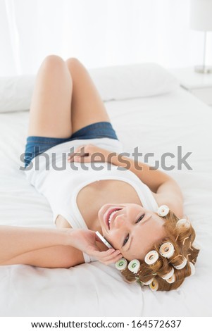 Portrait of a relaxed young woman in hair curlers using cellphone while lying in bed at home