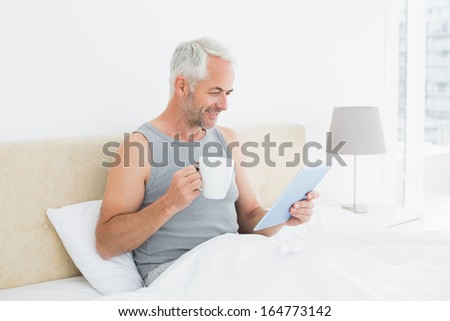 Side view of a smiling mature man with digital tablet and coffee table sitting in bed at home