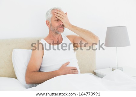 Thoughtful mature man sitting in bed with hand over closed eyes at home