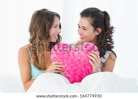 Two smiling young female friends with heart shaped pillow sitting in bed at home