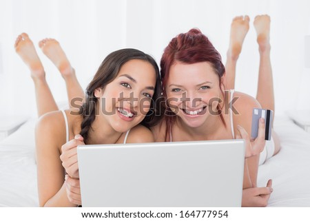 Portrait of happy relaxed young female friends doing online shopping through laptop and credit card in bed