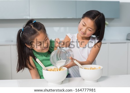 Two happy young girls pouring milk in bowl in the kitchen at home