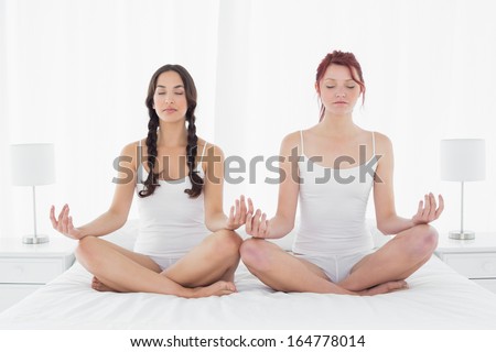 Full length of two young women with eyes closed sitting in lotus posture on bed at home